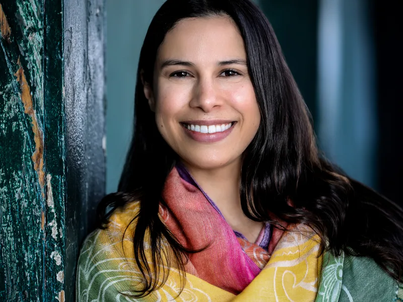 La Dra. Corina Lopez sonriendo, con una bufanda estampada en amarillo y rosa frente a una pared verde azulada desgastada